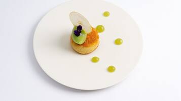 Un plato blanco con un postre que incluye una base redonda pequeña cubierta con una porción de fruta, una cucharada de crema verde y unas escamas de caviar.