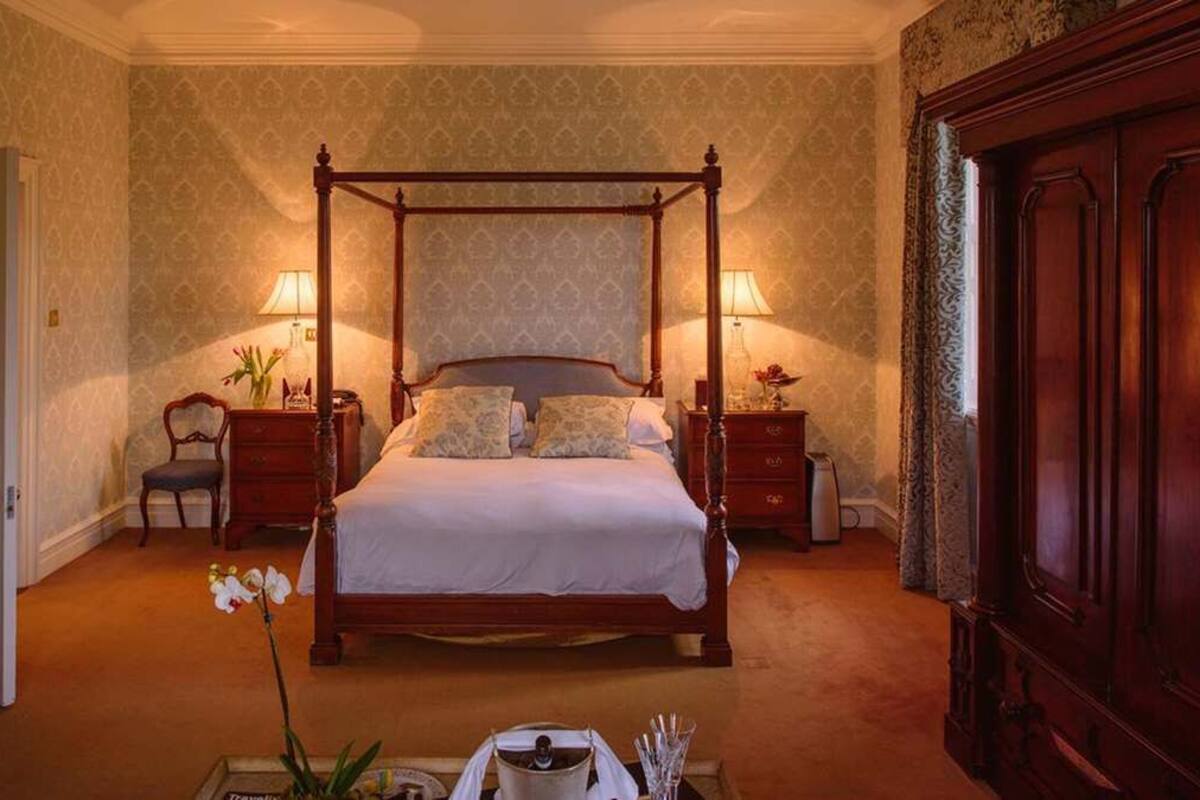 A four-poster bed with a canopy, two bedside tables with lamps, a chair, a dresser, and a vase with flowers.