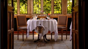 A dining area with a table set for four, surrounded by chairs, with a view of greenery through large windows.