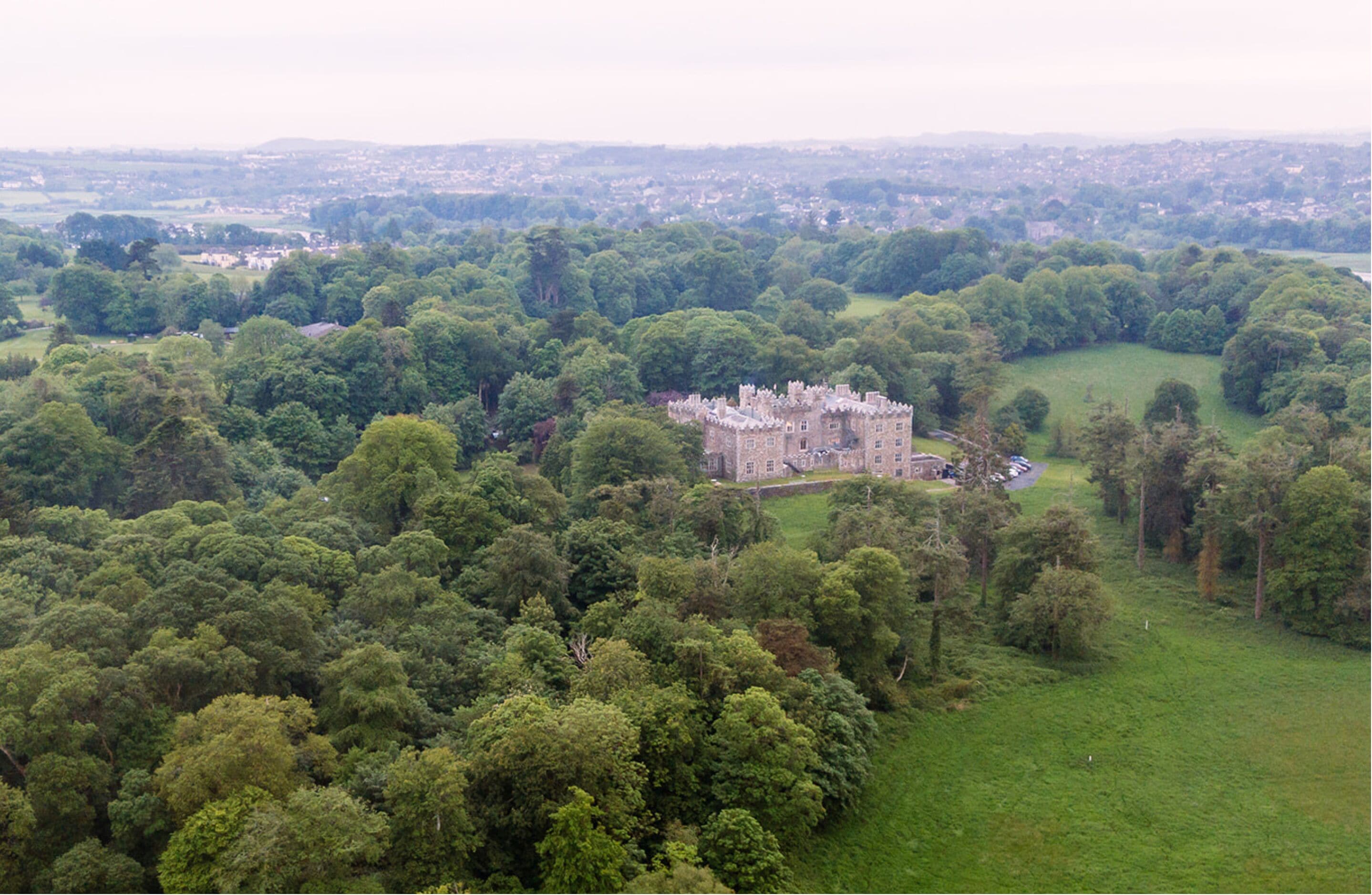 A large, historic building surrounded by dense greenery, with a well-maintained lawn and a distant view of a town.