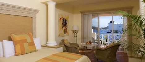 A hotel room with a bed, a chair, a small table with fruit, a lamp, and a view of the sea and buildings.