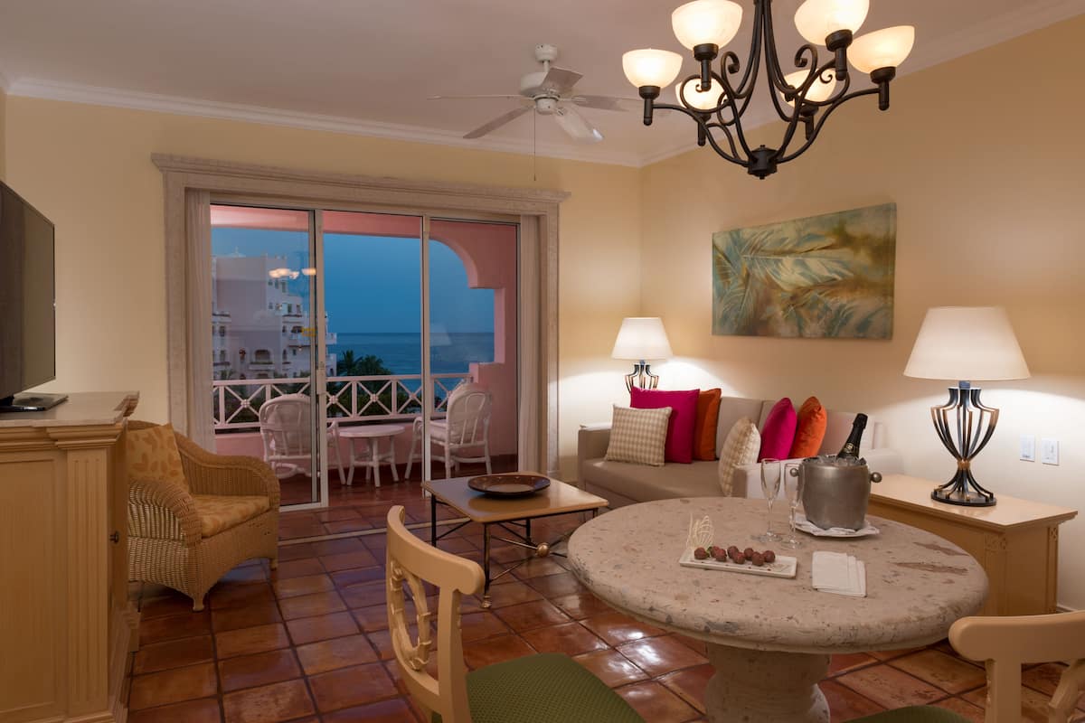 A living room with a balcony, a ceiling fan, a chandelier, a table with a bottle and glasses, and a painting on the wall.