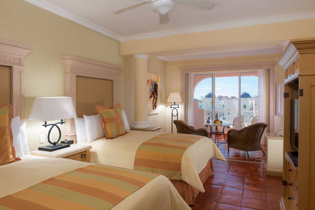 A hotel room with two beds, a TV, a dining area with a table and chairs, and a view of the city.