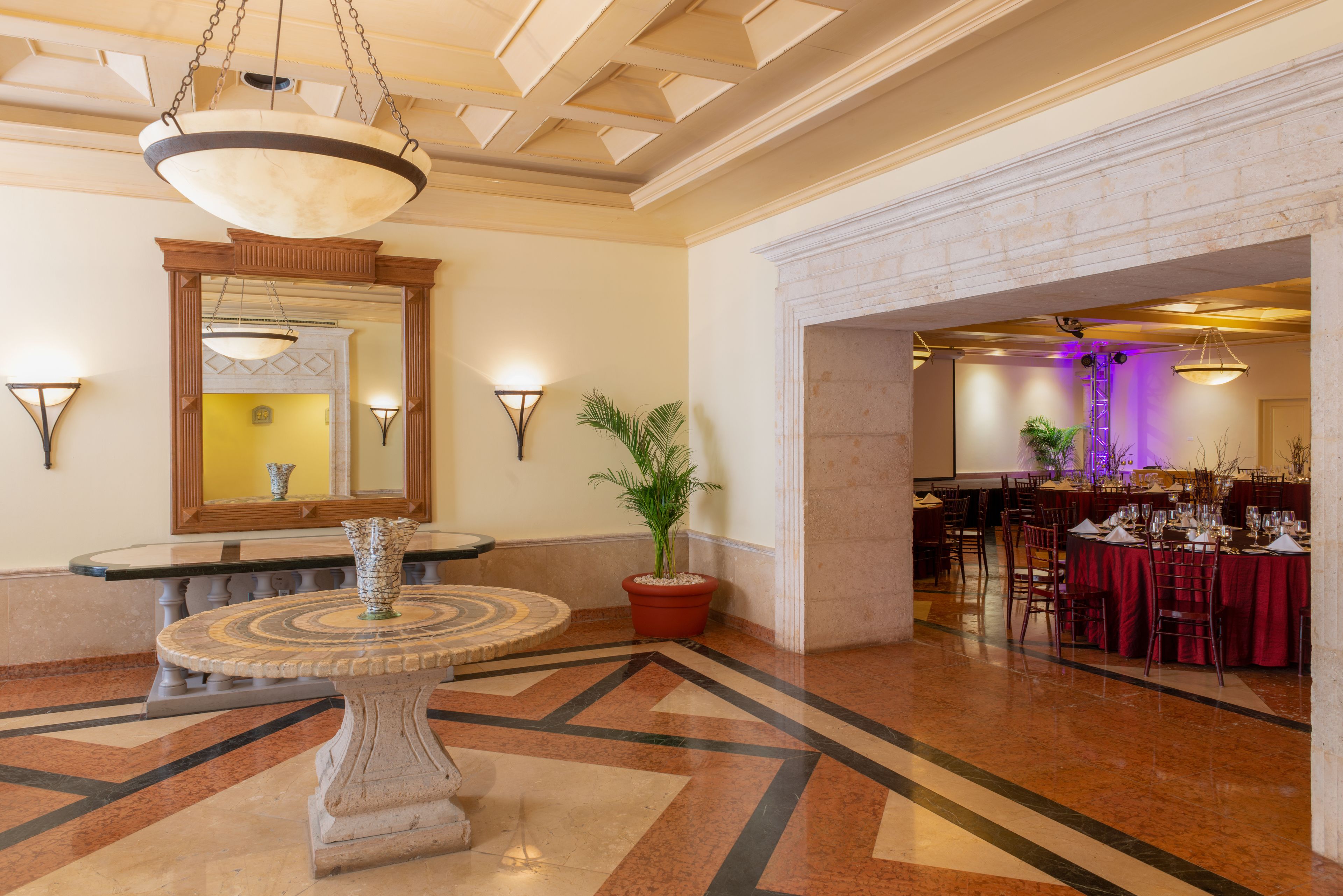 A lobby with a marble floor, a round table with a vase, a potted plant, and a dining area with tables set for a meal.