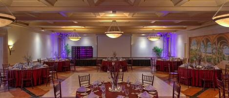 Un salón de banquetes con mesas redondas dispuestas para el servicio de comidas, con una araña central e iluminación ambiental.
