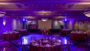 Une salle de banquet avec des tables rondes dressées pour le repas, un grand écran et une fresque murale.
