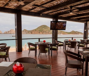 Un espacio exterior para comer con mesas de madera y sillas de mimbre, con vistas a un paisaje costero de colinas rocosas y aguas tranquilas.