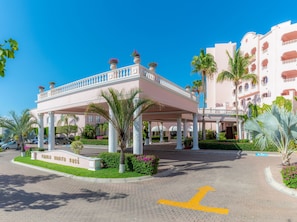 Entrada de hotel con porche cubierto, palmeras y cielo azul despejado.