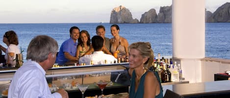 A group of people dining at an outdoor bar with a view of the sea and rocky cliffs.