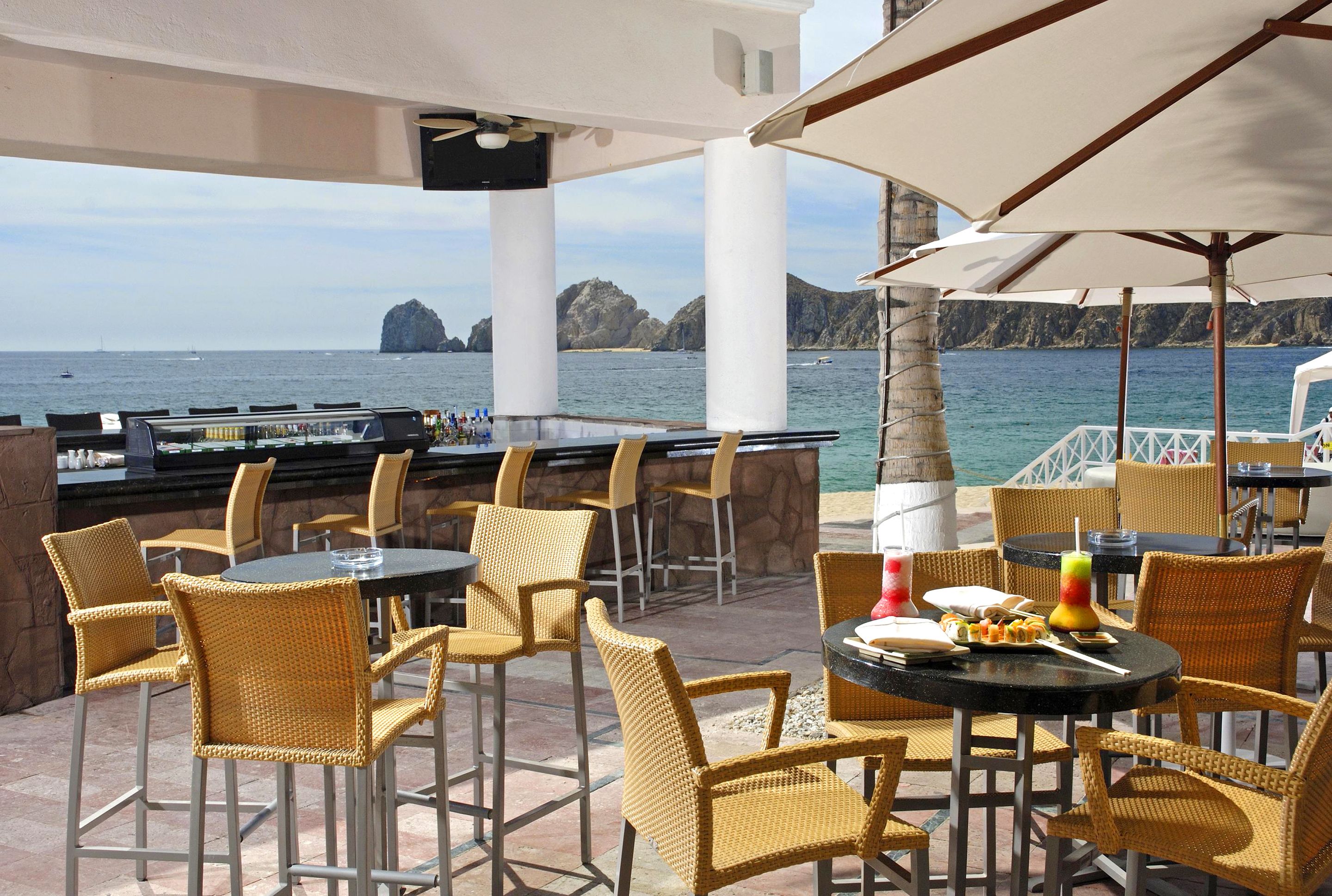Zona de bar al aire libre con sillas y mesas de mimbre, con vistas al mar y acantilados rocosos al fondo.