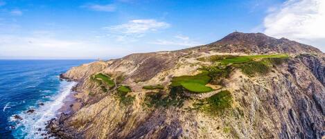 A rugged coastline with a mountain, a golf course, and a small building.