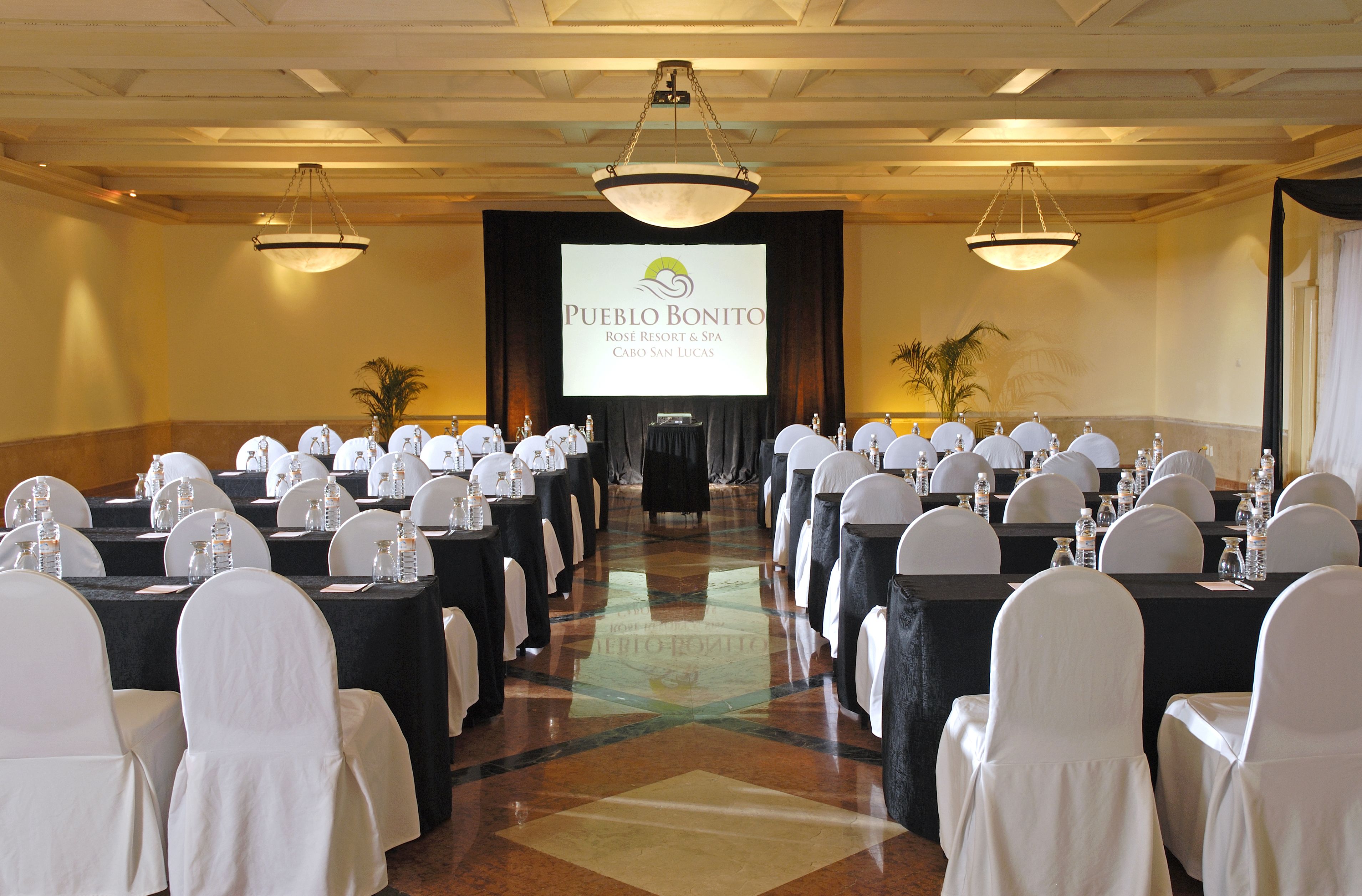 A conference room with rows of tables and chairs, a large screen displaying a presentation, and a chandelier.
