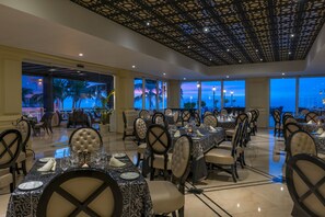 A dining area with tables set for service, large windows with a view of palm trees and a sunset, and a decorative ceiling.