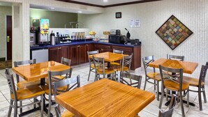 A hotel dining area with wooden tables and chairs, a buffet counter with various food items, and a wall-mounted quilt. - Country Hearth Inn Knightdale Raleigh (Knightdale)