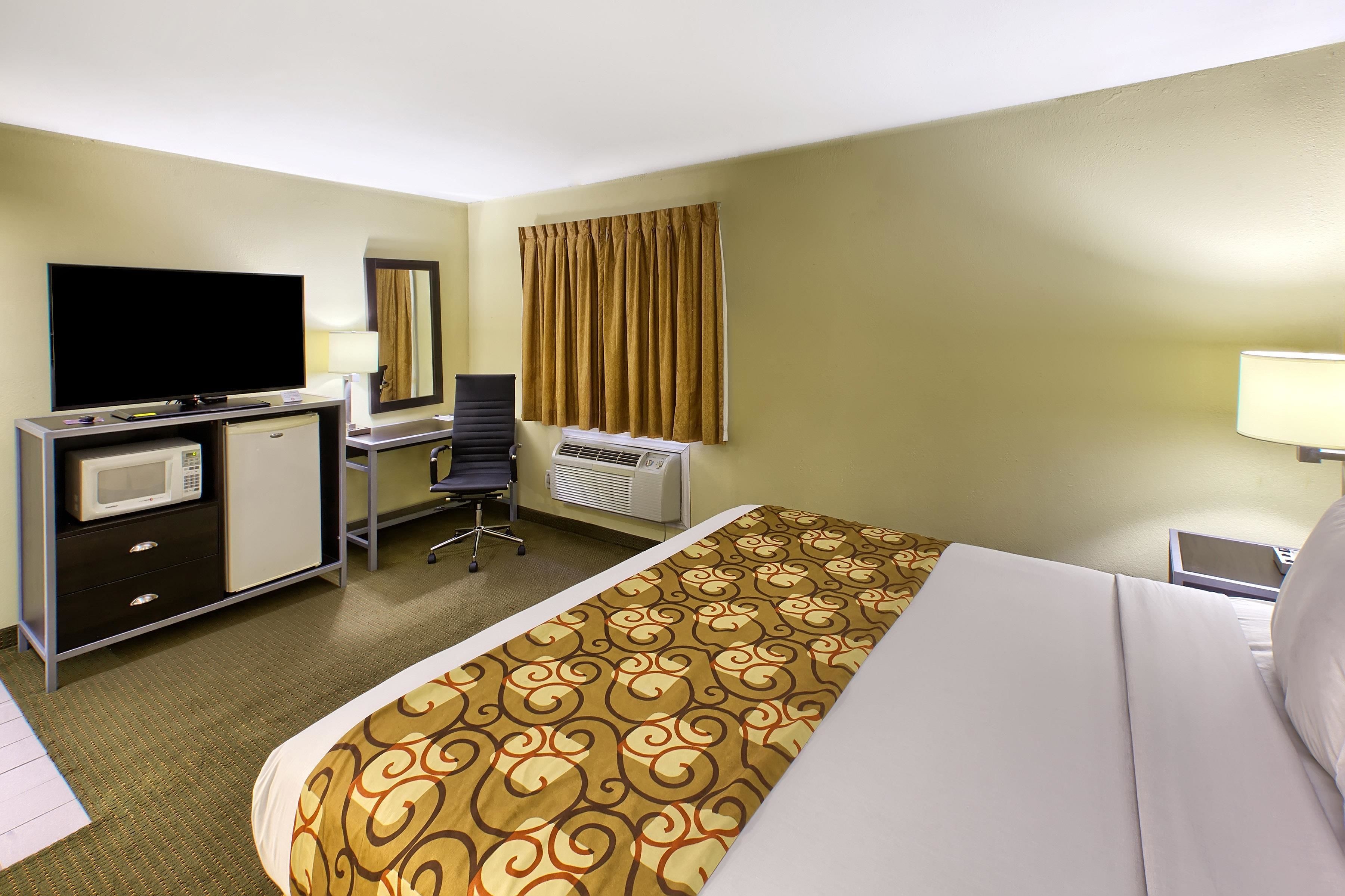 A hotel room with a bed, a desk, a television, a microwave, a chair, and an air conditioning unit.