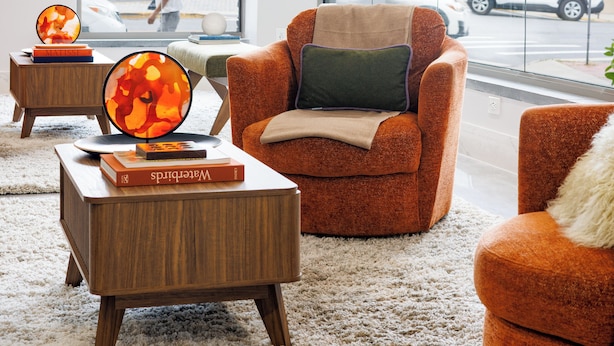 Un vestíbulo de hotel moderno con un gran ventanal, un sillón naranja cómodo, una mesita de madera con libros y una mesa redonda con objetos decorativos.