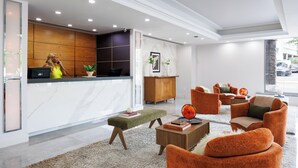 Un área de recepción moderna con mostrador de mármol, paneles de madera y una zona de estar con sillas naranjas y un manto de alfombra blanca esponjosa.