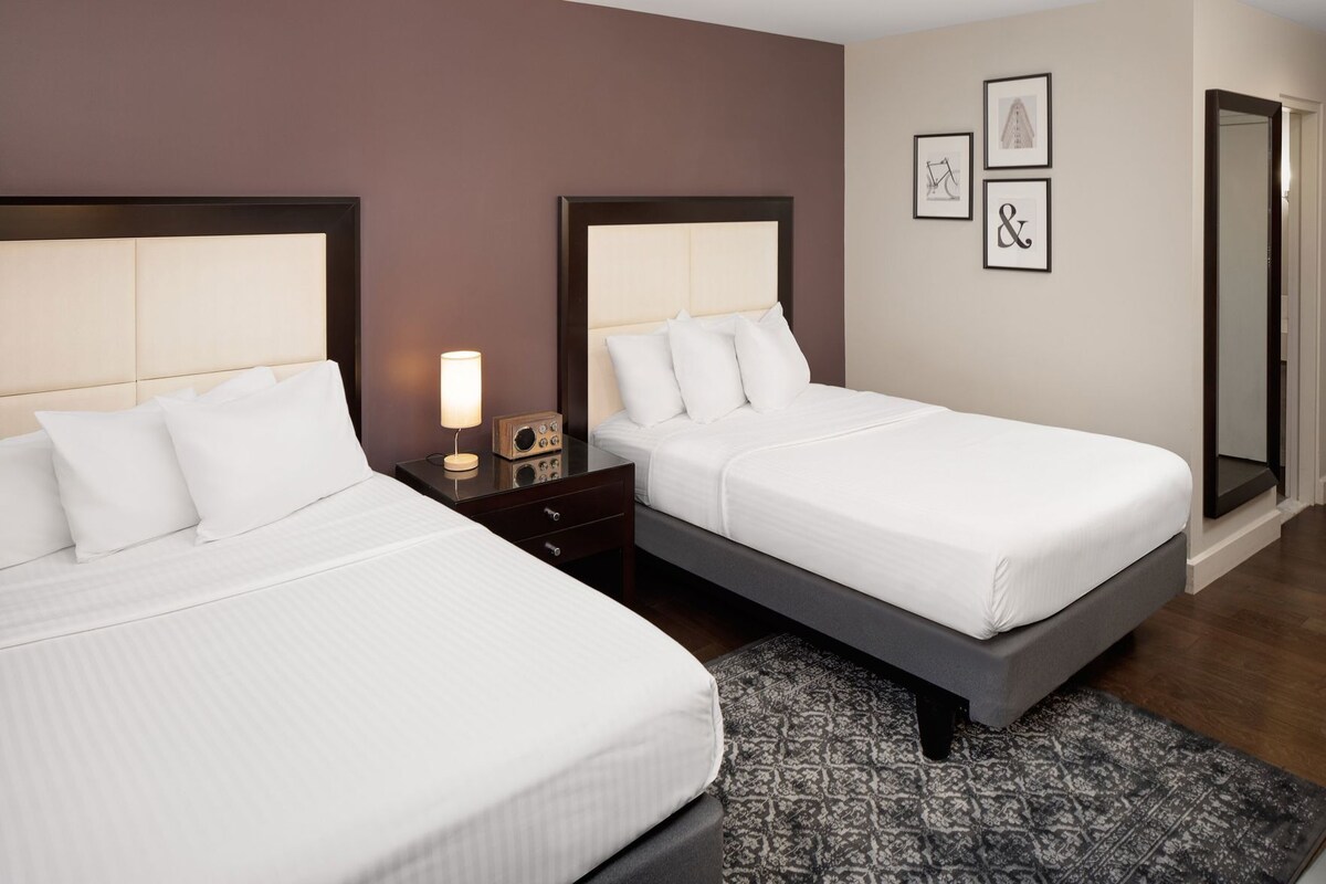 A hotel room with two beds, a bedside table with a lamp, a patterned rug, and framed artwork on the wall.