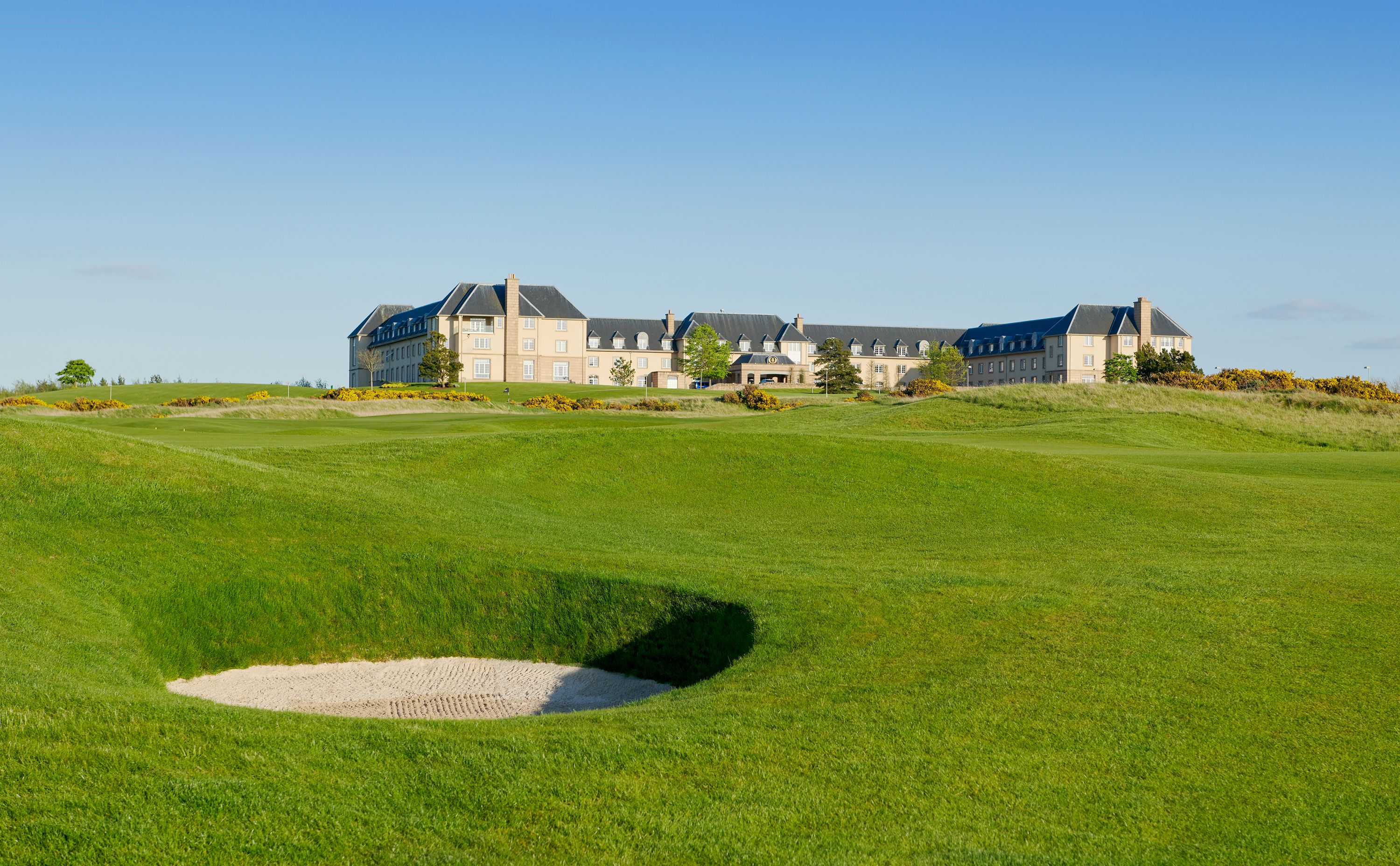 Un grand bâtiment à étages, doté d’une façade symétrique, situé sur une pelouse bien entretenue, avec un bunker de sable en premier plan.