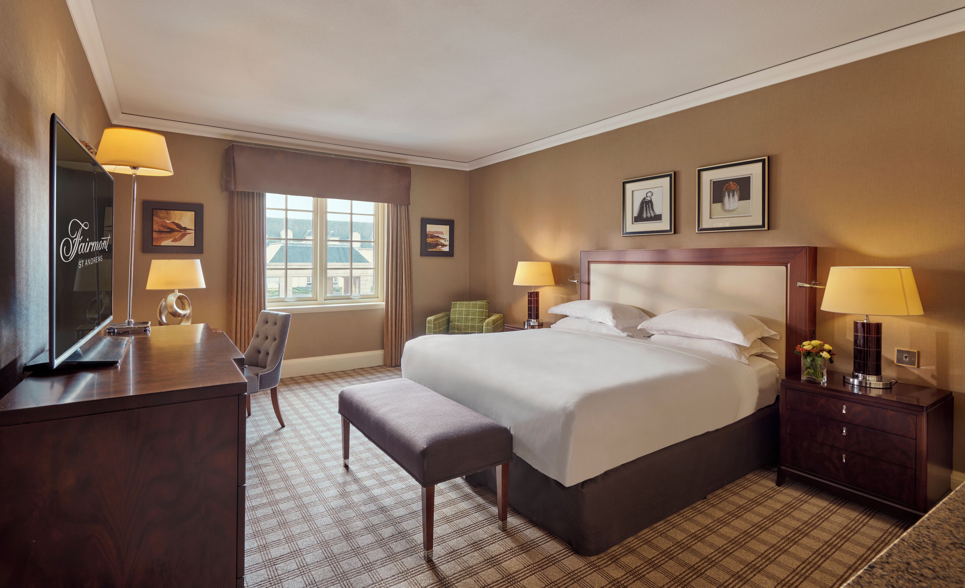 A hotel room with a large bed, a desk with a TV, a chair, a bedside table with a lamp, and a window with curtains.