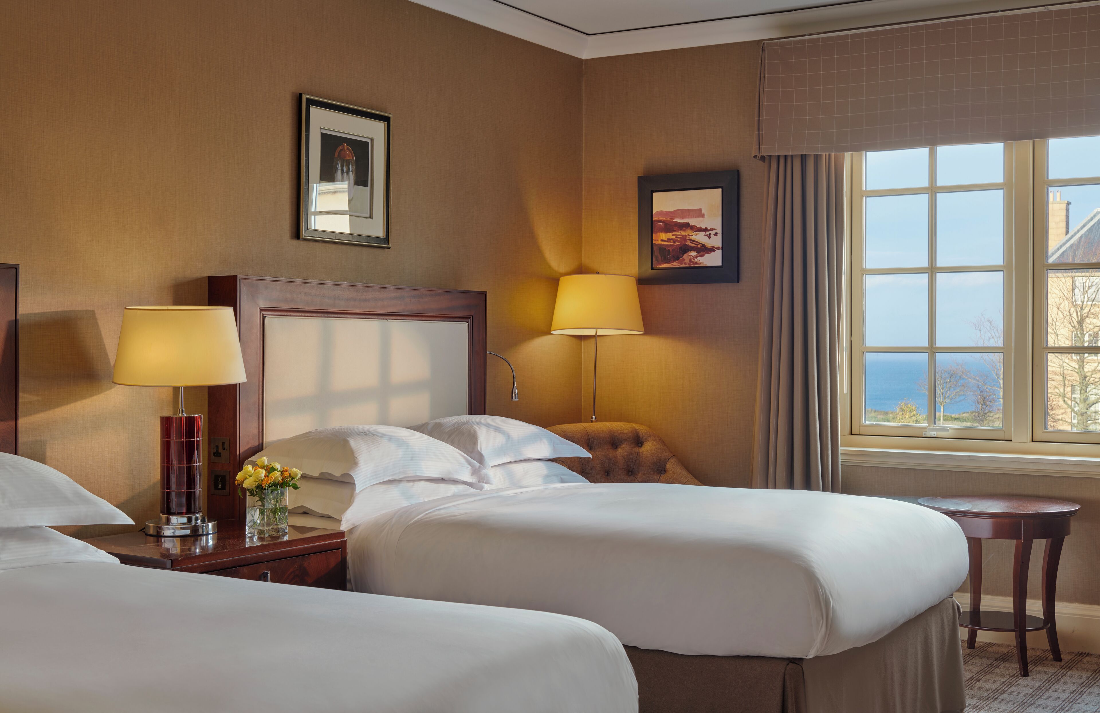 Habitación de hotel con dos camas, una mesita de noche con lámpara, una pequeña mesa y ventana con vista al mar.