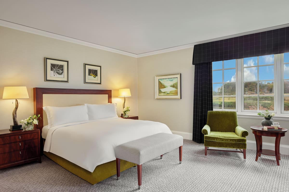 A hotel room with a large bed, two bedside tables with lamps, a green armchair, a small table with a vase, and two framed pictures on the wall.