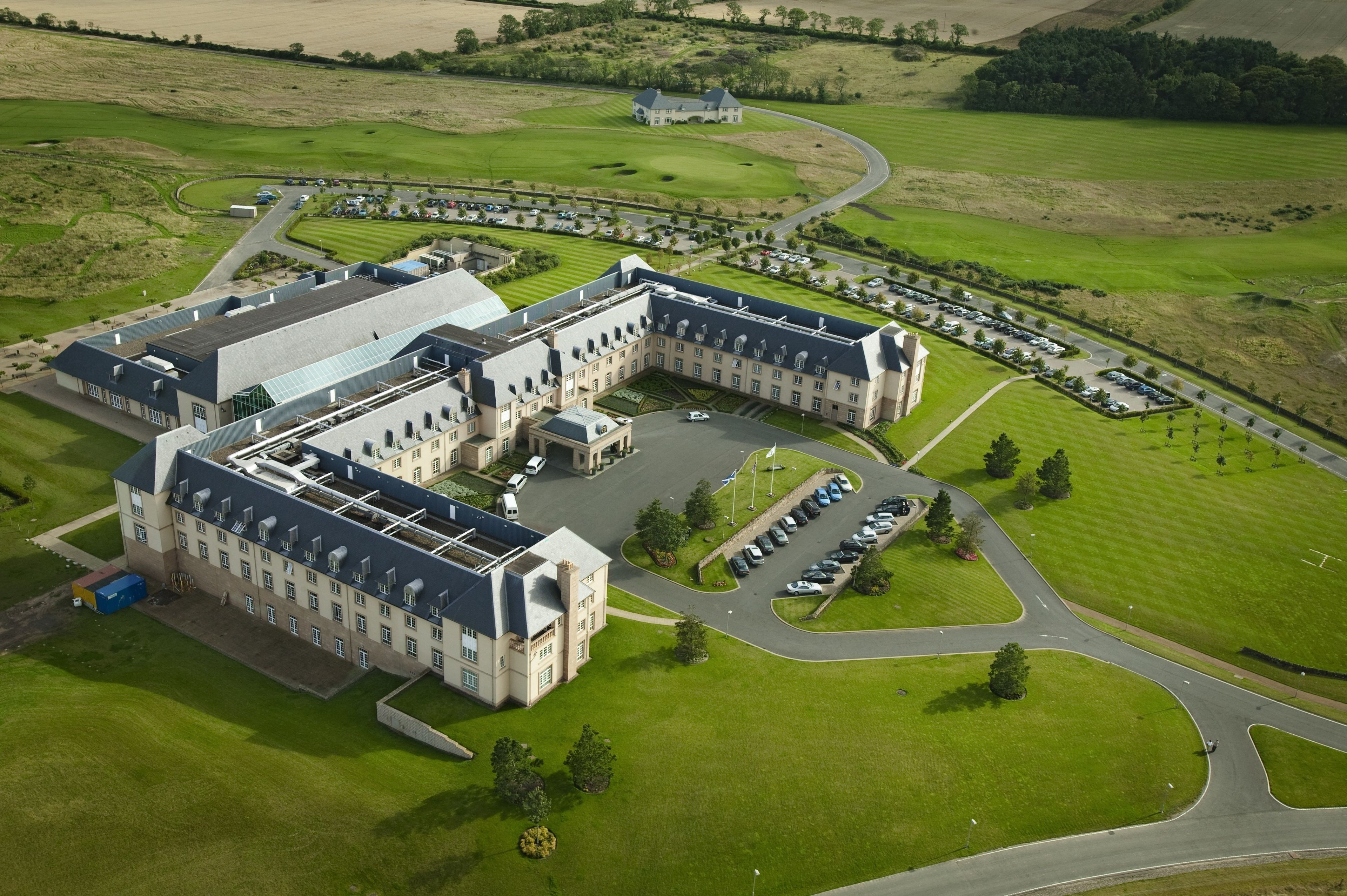 An aerial view of a large, L-shaped building with multiple wings, surrounded by well-maintained lawns and a golf course.