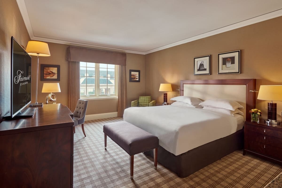 A hotel room with a large bed, a desk, a chair, and a TV. There are two windows with curtains, a nightstand with a lamp, and two framed pictures on the wall.