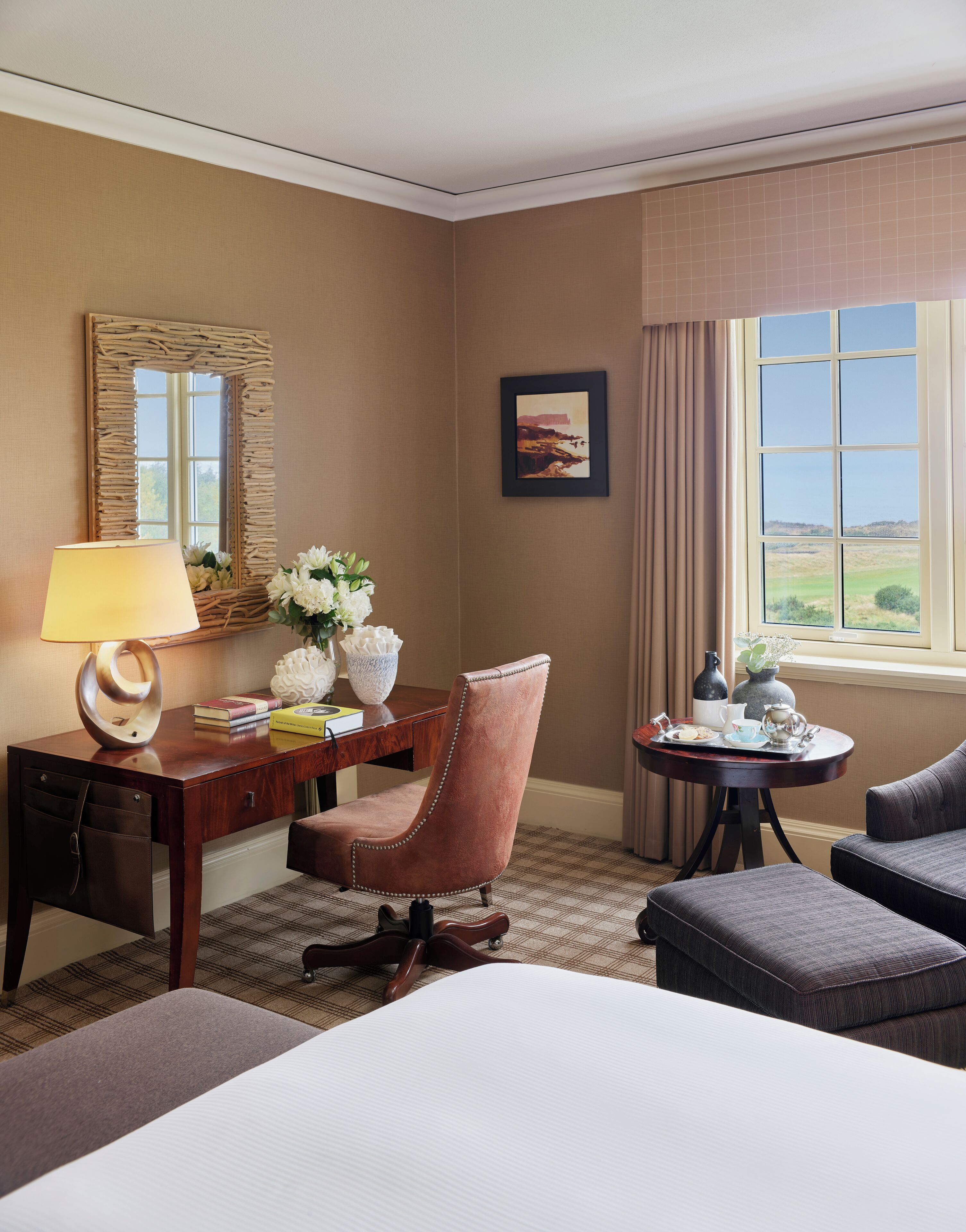 Ein Hotelzimmer mit Bett, Schreibtisch, Stuhl, Tisch mit Tablett, Lampe, Spiegel, Bild und Fenster mit Aussicht.