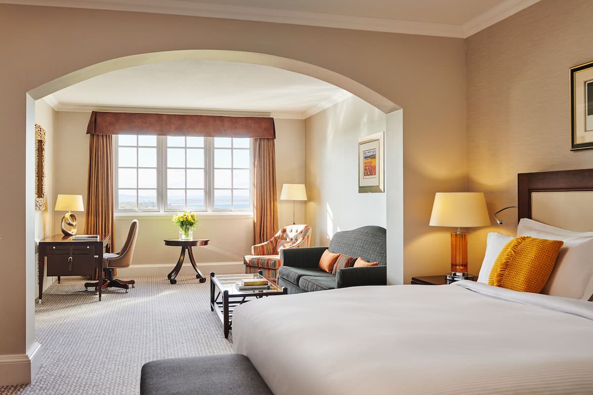 A hotel room with a large bed, a desk, two chairs, a small table with a lamp, and a window with curtains.