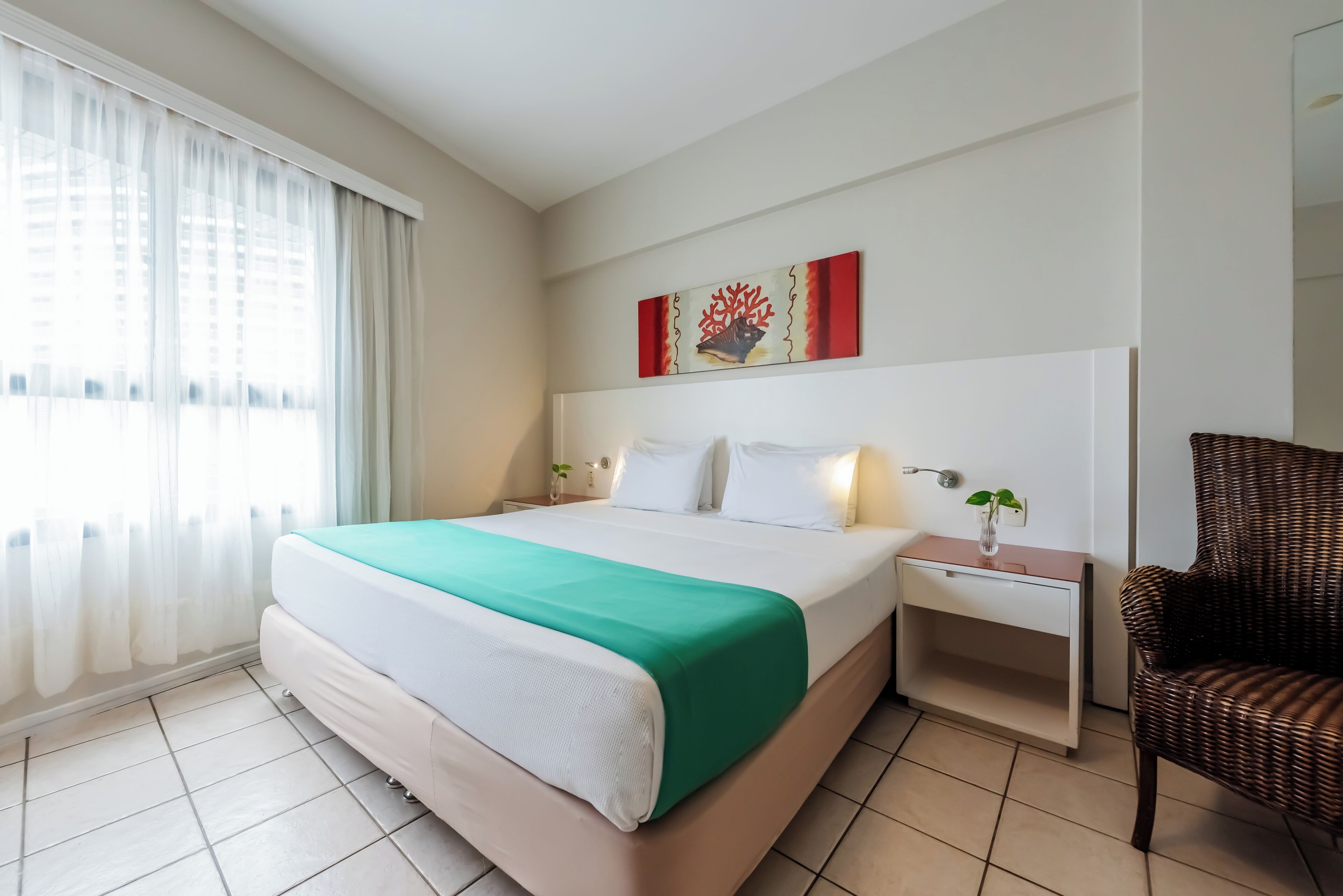 Une chambre d’hôtel avec un grand lit, une table de chevet ornée d’un vase, un fauteuil en osier et un tableau accroché au mur.