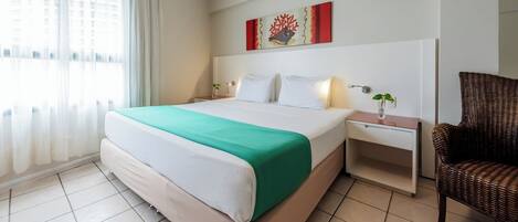 A hotel room with a large bed, a bedside table with a vase, a wicker chair, and a painting on the wall.