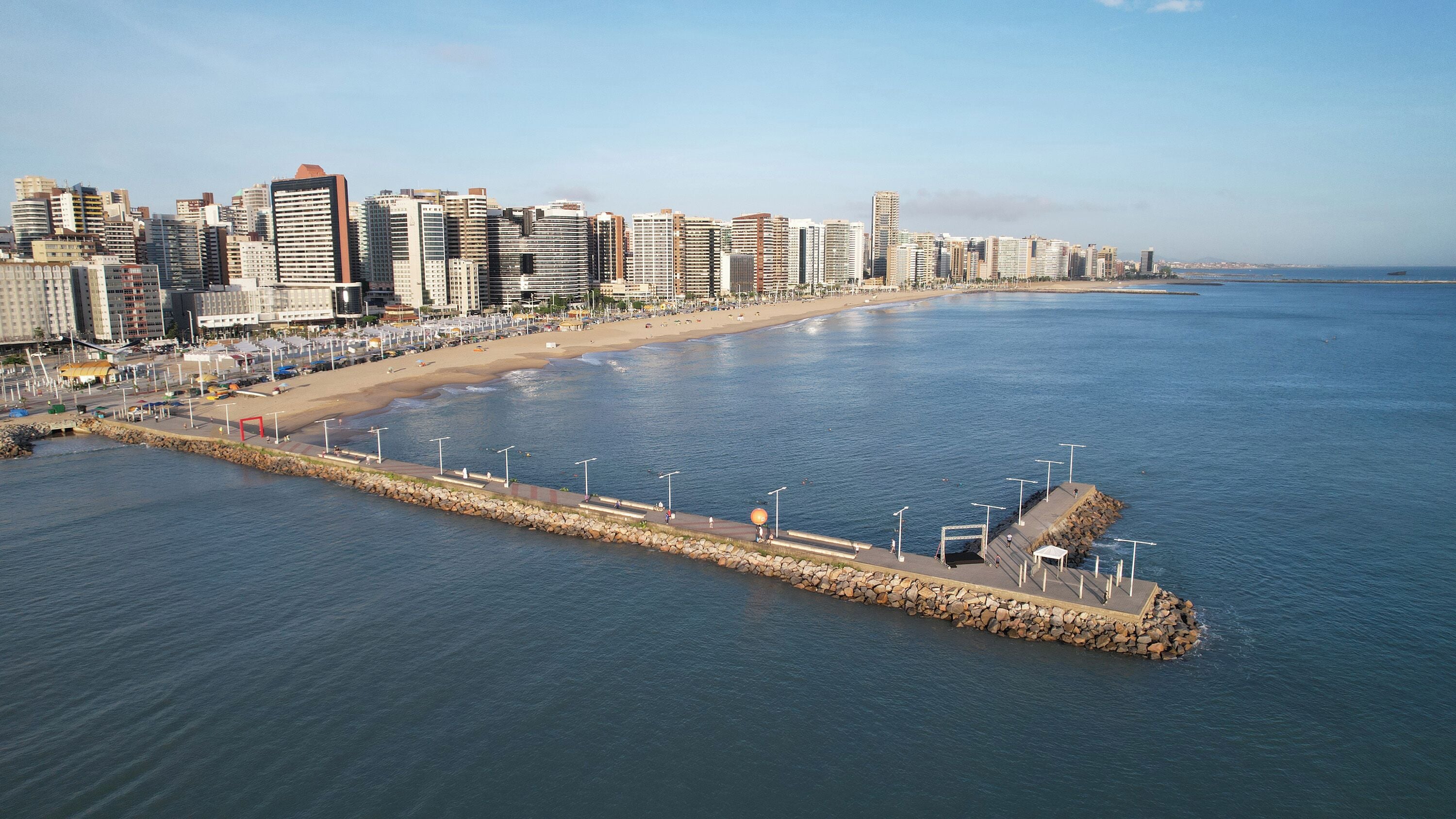 A coastal cityscape with a wide beach, a stone pier, and high-rise buildings.