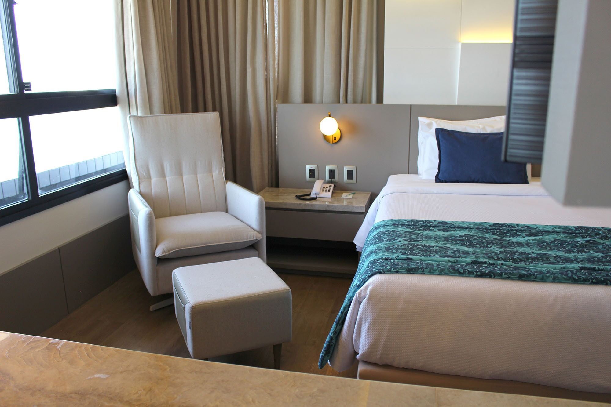 A hotel room with a bed, a chair, a nightstand with a phone, and a window with curtains.