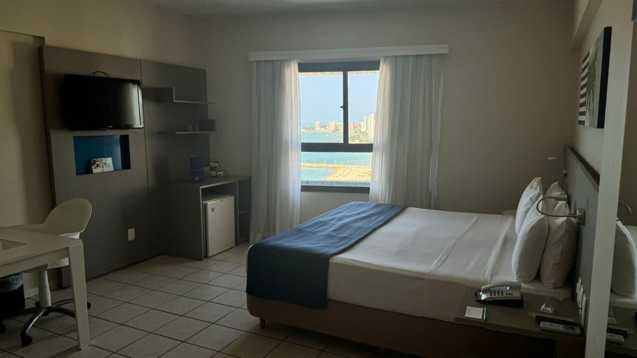 A hotel room with a bed, a desk, a television, and a view of the beach.