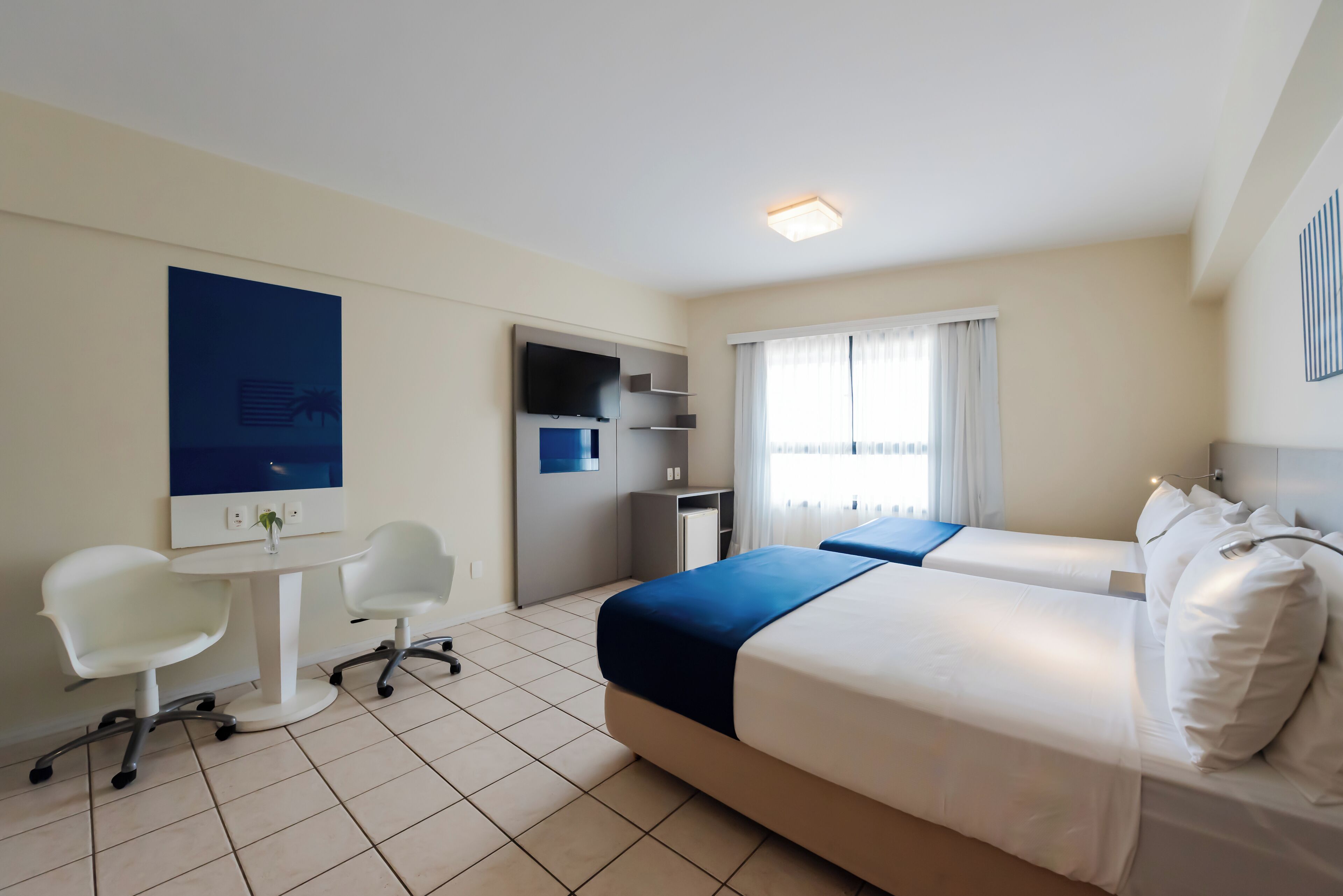 A hotel room with a bed, two white chairs, a small desk, a TV, and a window with curtains.