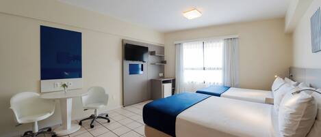 A hotel room with a bed, two white chairs, a small desk, a TV, and a window with curtains.