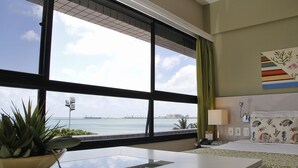 Habitación de hotel con un ventanal que ofrece vistas al mar y al horizonte urbano, una mesita de noche con una planta, una cama con almohada estampada y una lámpara de noche.
