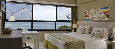 Habitación de hotel con un ventanal grande, una cama con ropa de cama estampada, un escritorio con lámpara y un cuadro en la pared.