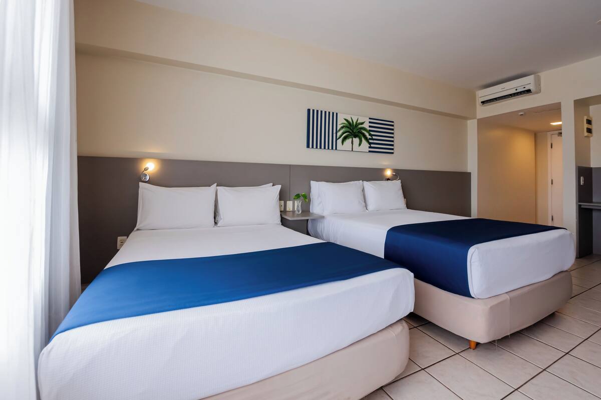 Twee tweepersoonsbedden met wit en blauw beddengoed, een wandairconditioner en een kleine tafel met een plant.