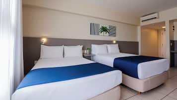 Two double beds with white and blue bedding, a wall-mounted air conditioner, and a small table with a plant.