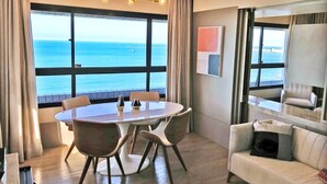 Habitación de hotel moderna con un ventanal grande que ofrece vistas al mar, una mesa redonda para comer con sillas, un sofá blanco y un televisor pequeño.
