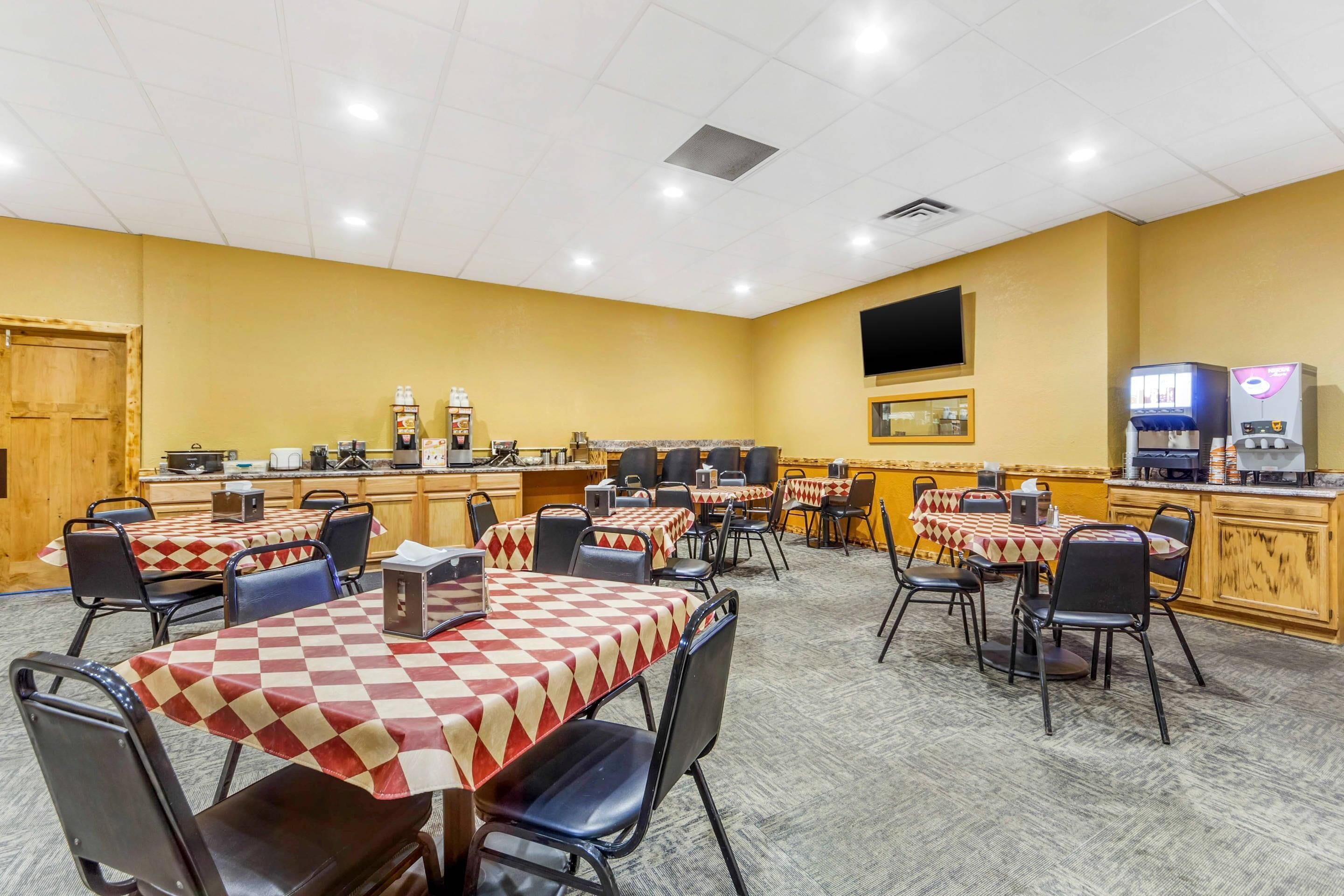 A hotel dining area with multiple tables and chairs, a kitchen area, and a television mounted on the wall.