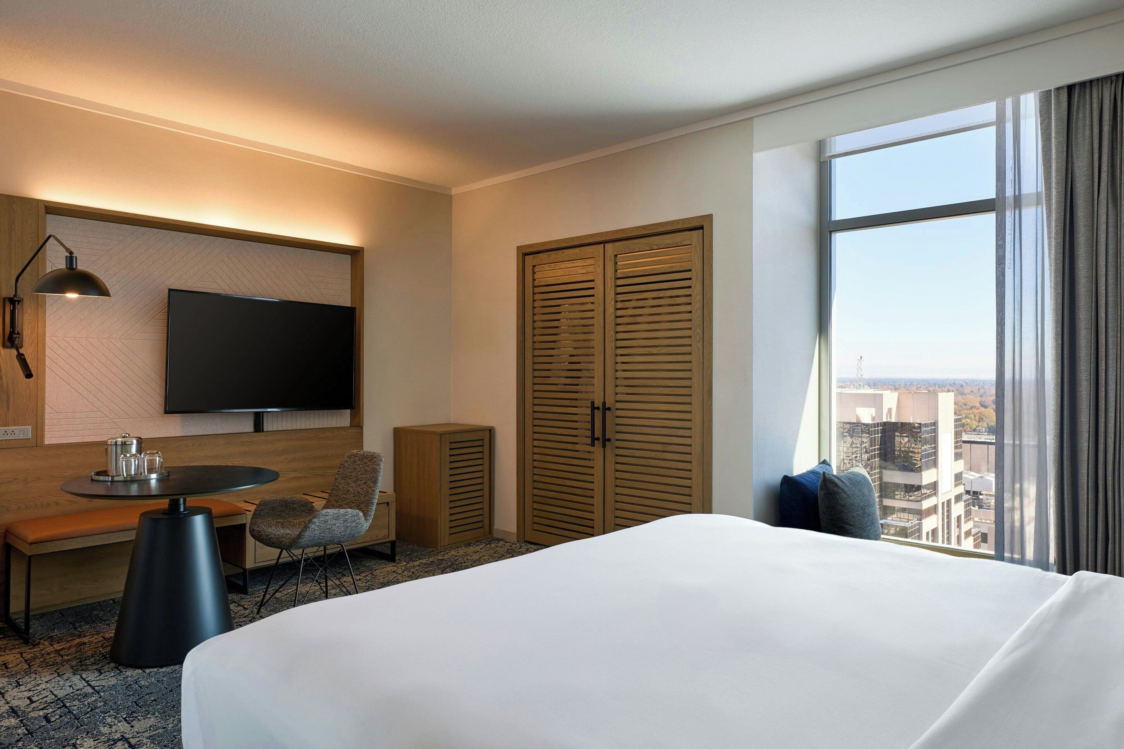 Habitación de hotel con una cama grande, un televisor montado en la pared, un escritorio con una silla, una mesita de noche con una lámpara y un armario.