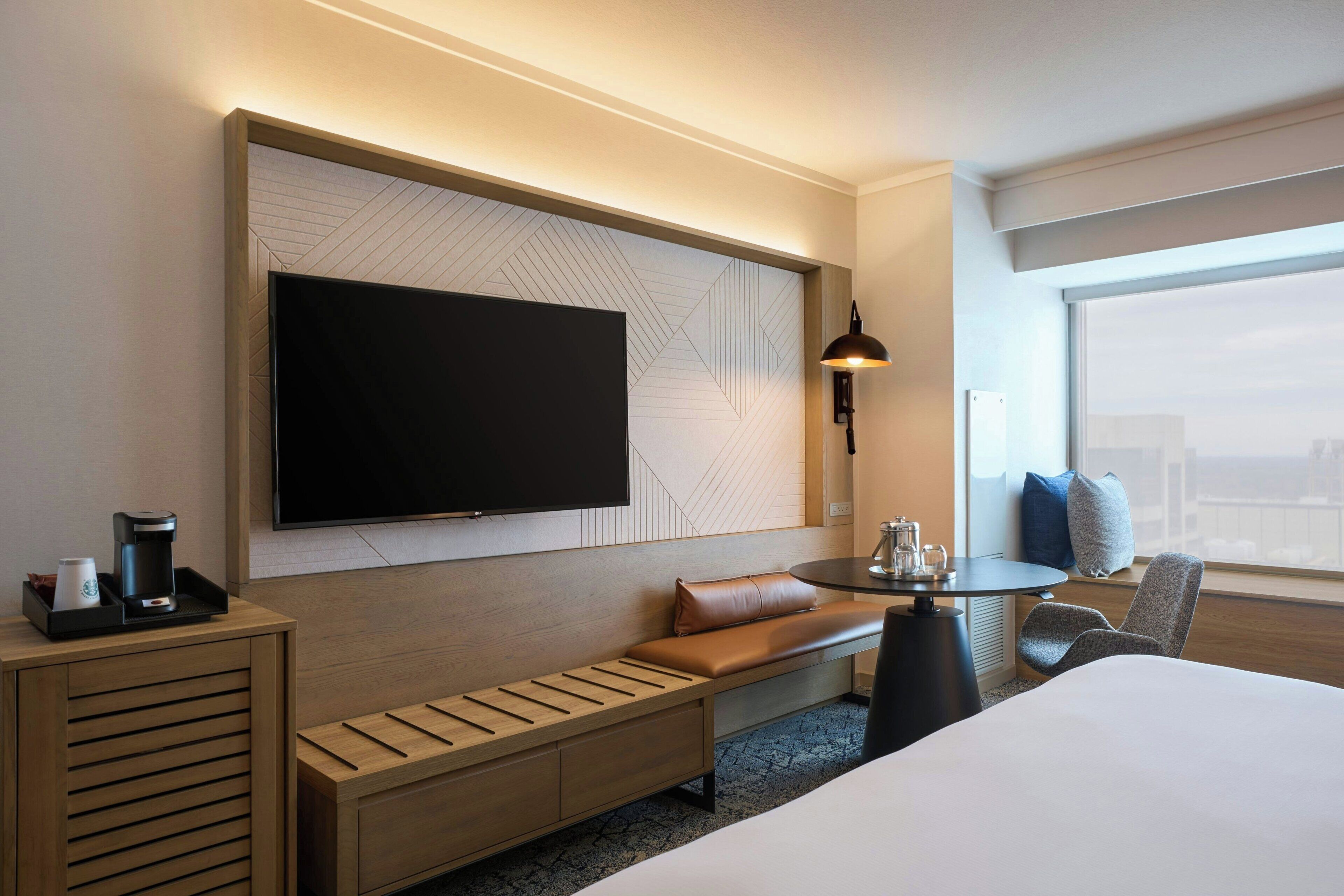 A hotel room with a large flat-screen TV mounted on the wall, a wooden bench with storage, a round table with a lamp, and a large window offering a city view.