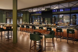A bar area with wooden flooring, green bar stools, and a variety of bottles and glasses on the bar counter.