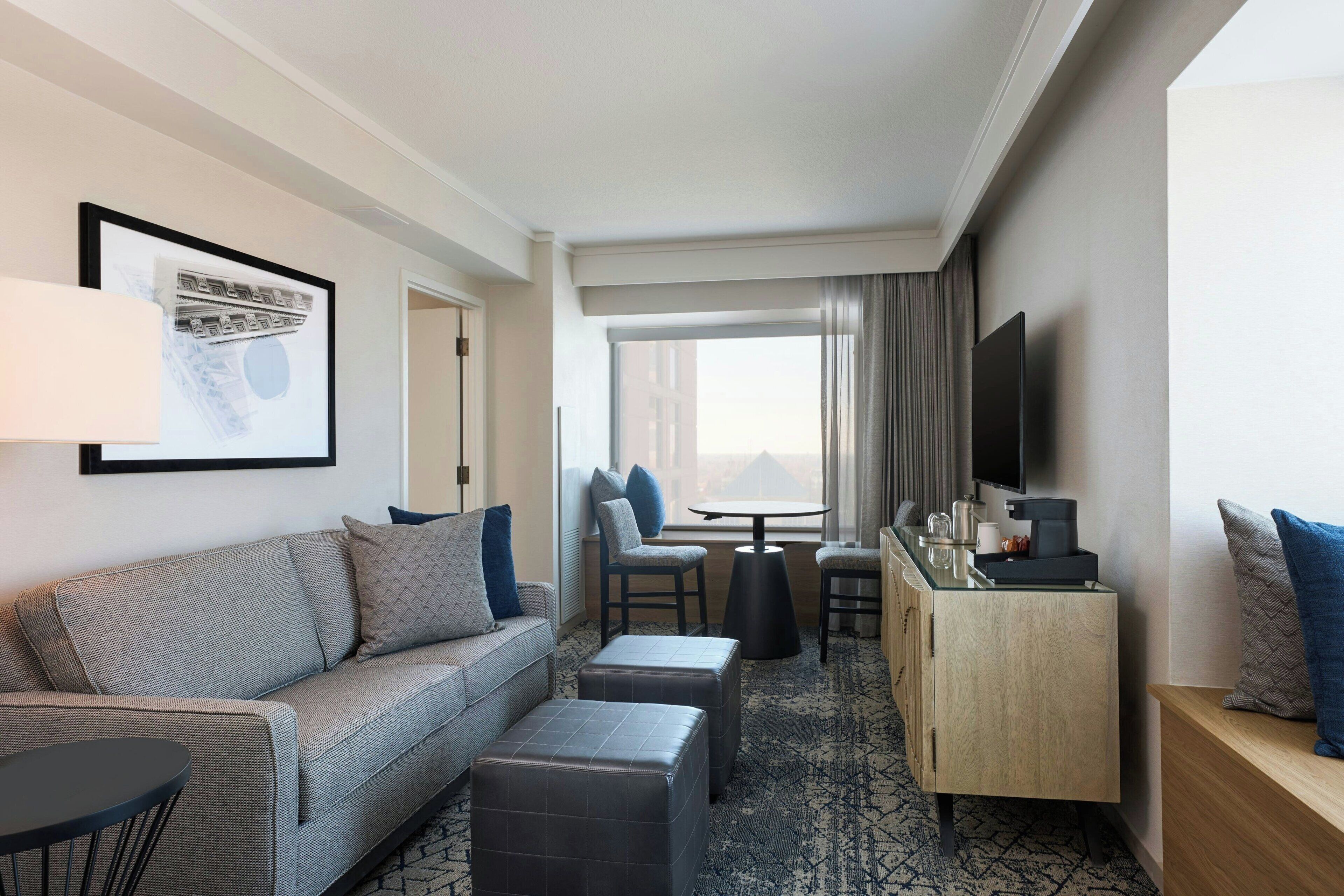 Ein Hotelzimmer mit einer Couch, einem Hocker, einem kleinen Tisch, einem Fernseher und einem Schreibtisch mit Stuhl.
