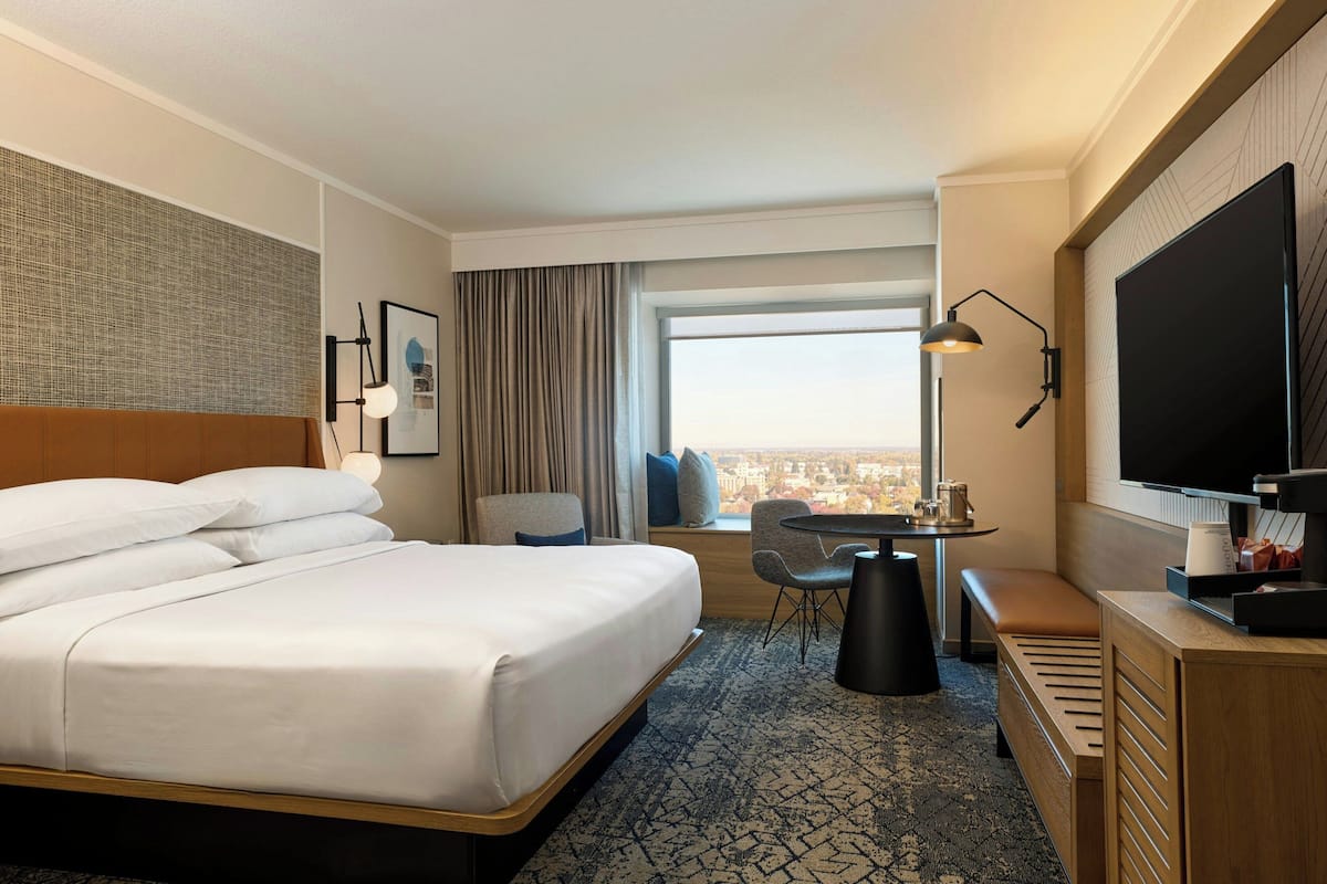 Ein Hotelzimmer mit einem großen Bett, einem Schreibtisch mit Stuhl, einem Fernseher und einem Fenster mit Blick auf die Stadt.