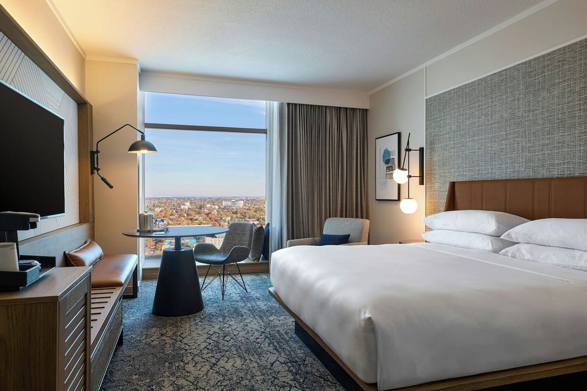 Ein Hotelzimmer mit einem großen Bett, einem Schreibtisch mit Stuhl, einem kleinen Tisch und Blick auf die Stadt durch ein großes Fenster.