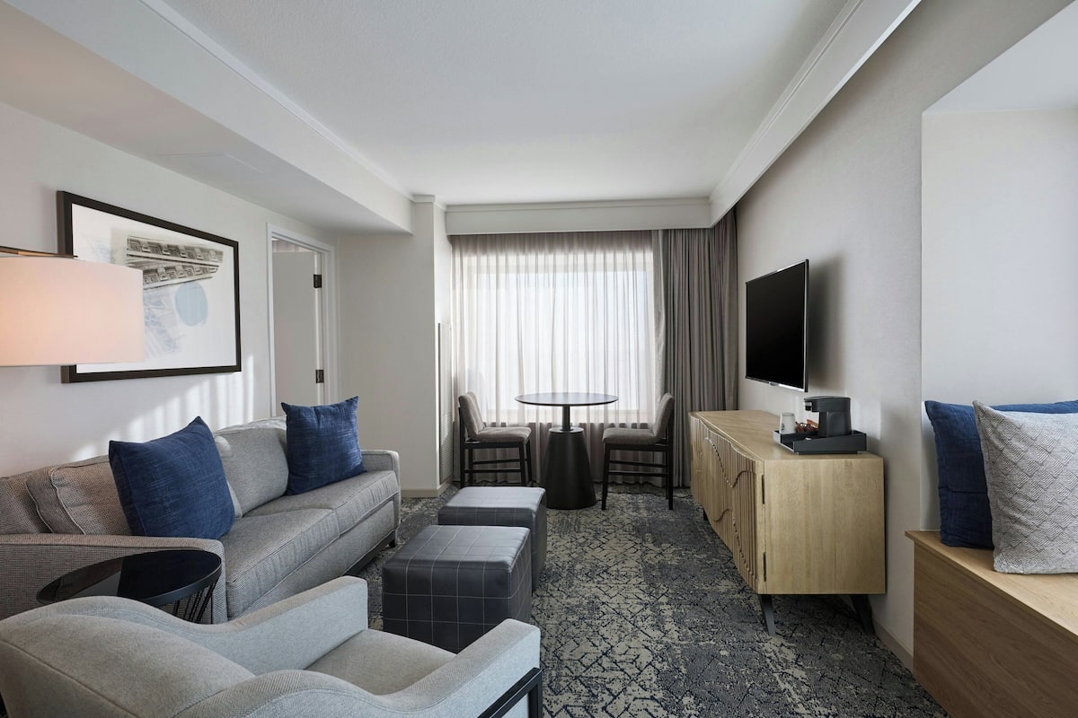 Ein modernes Hotelzimmer mit grauem Sofa, Poufs, einem kleinen Tisch, einem Fernseher und einem Holzschrank.